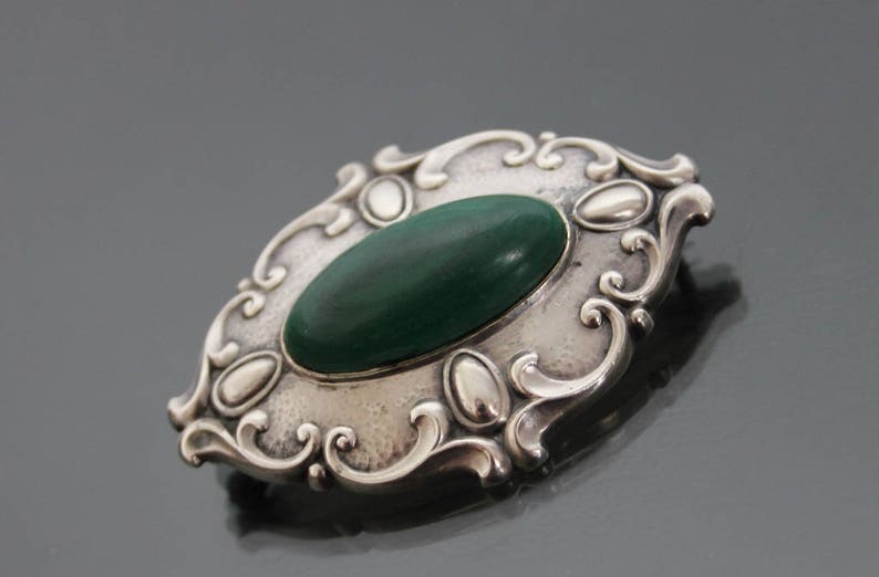 May include: A silver brooch with an oval-shaped green stone set in a decorative frame.