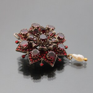 Op de afbeelding: Een rode granaat broche met een parel hanger. De broche heeft de vorm van een bloem met een stervormig ontwerp.
