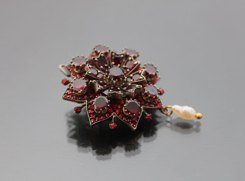Op de afbeelding: Een rode granaat broche met een parel hanger. De broche heeft de vorm van een bloem met een stervormig ontwerp.