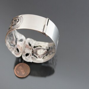 May include: A silver bracelet with a hinged clasp and intricate cut-out designs. The bracelet is open and lying on a gray surface with a penny underneath.
