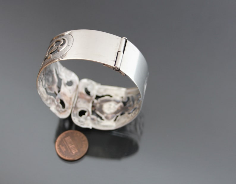 May include: A silver bracelet with a hinged clasp and intricate cut-out designs. The bracelet is open and lying on a gray surface with a penny underneath.