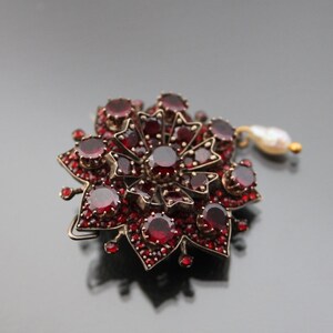 Op de afbeelding: Een rode granaat broche in de vorm van een bloem met een parel hanger.