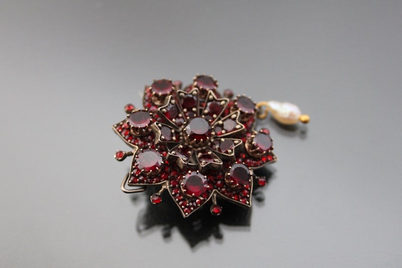 Op de afbeelding: Een rode granaat broche in de vorm van een bloem met een parel hanger.