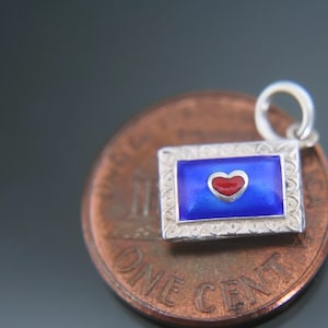 May include: A small silver charm in the shape of a blue and white envelope with a red heart inside. The charm is sitting on top of a US penny.