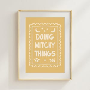 May include: A framed wall art print featuring a mustard yellow background with a white scalloped border. Inside the border, the text reads DOING WITCHY THINGS in white, accompanied by white illustrations of two crescent moons, stars, and leaves.