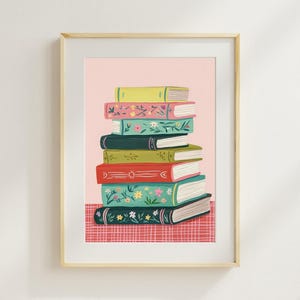 May include: A framed art print featuring a stack of colorful books with floral designs. The books have various colors, including green, red, and blue, and are set against a pink background. The text "Kind of Fine Studio" is at the bottom.