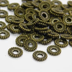 以下が含まれることがあります： ビーズの縁取りが施されたアンティークゴールド調の金属製スペーサービーズが大量に積み重ねられています。ビーズは丸型で、中央に穴が開いています。
