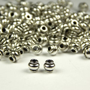 May include: A close-up of two silver metal beads with a textured design. The beads are round and have a small hole in the center. The beads are surrounded by a pile of similar beads.