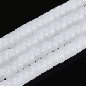 May include: Close-up of several strands of white, square-shaped beads. The beads are strung together, creating a textured, ribbed appearance. The beads have a glossy finish and reflect light, creating highlights. The background is black.