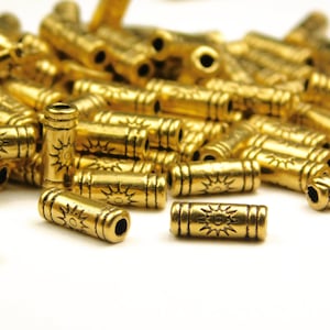 May include: Close-up of numerous gold-colored metal beads. Each cylindrical bead features a sunburst design and horizontal lines. The beads are scattered on a white surface, suggesting a craft or jewelry-making theme.