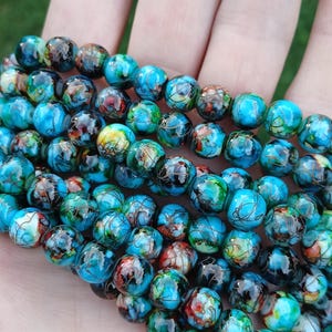 May include: Strands of colorful, round beads in shades of blue, green, red, and black. The beads have a marbled appearance and are held in a hand, suggesting they are for jewelry making or crafting.