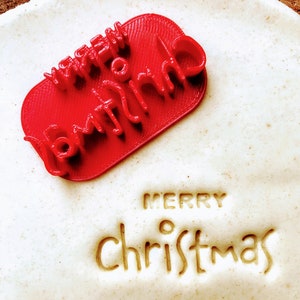 Può includere: Un timbro di plastica rosso con le parole "Merry Christmas" su di esso. Il timbro è su una superficie bianca con le parole "Merry Christmas" impresse nella superficie.