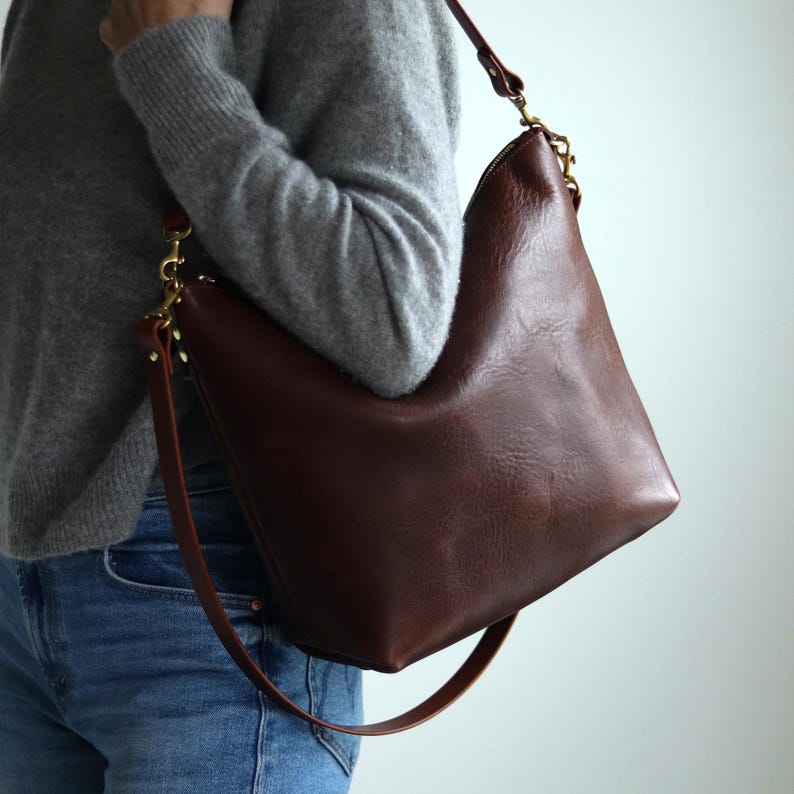 Puede incluir: Un bolso de hombro de cuero marr&oacute;n con una correa larga. El bolso tiene un aspecto informal y holgado y se lleva sobre el hombro de una persona que lleva vaqueros y un jersey gris.