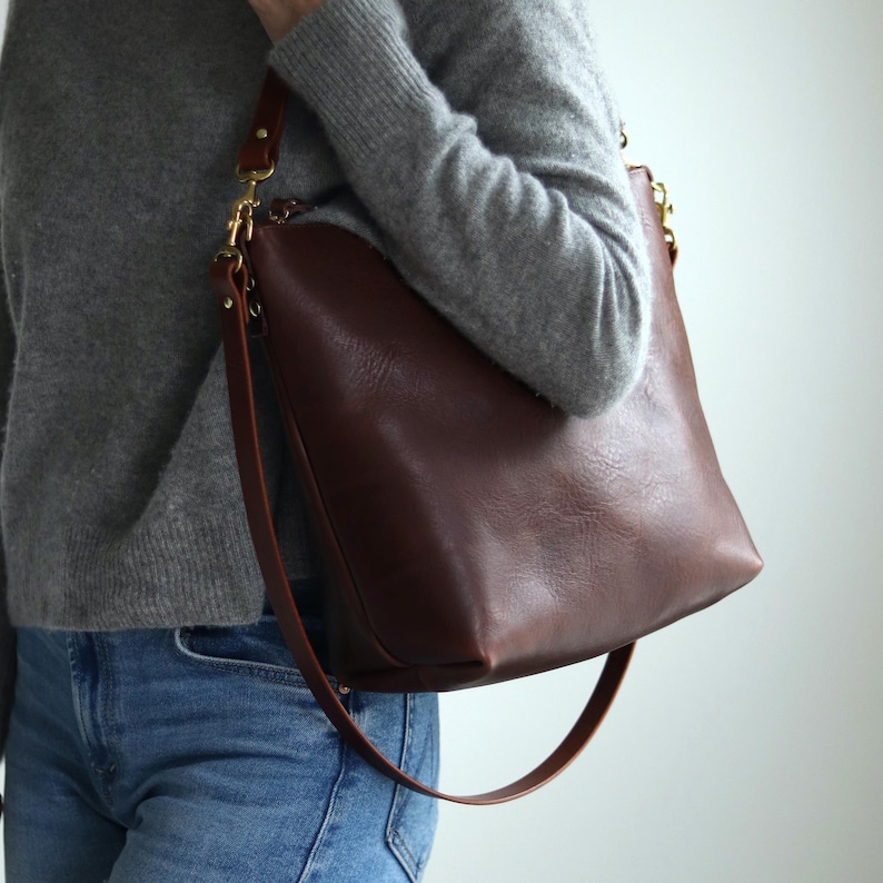 Puede incluir: Un bolso tote de cuero marr&oacute;n con una correa larga ajustable. El bolso tiene una cremallera superior y una hebilla de metal dorado.