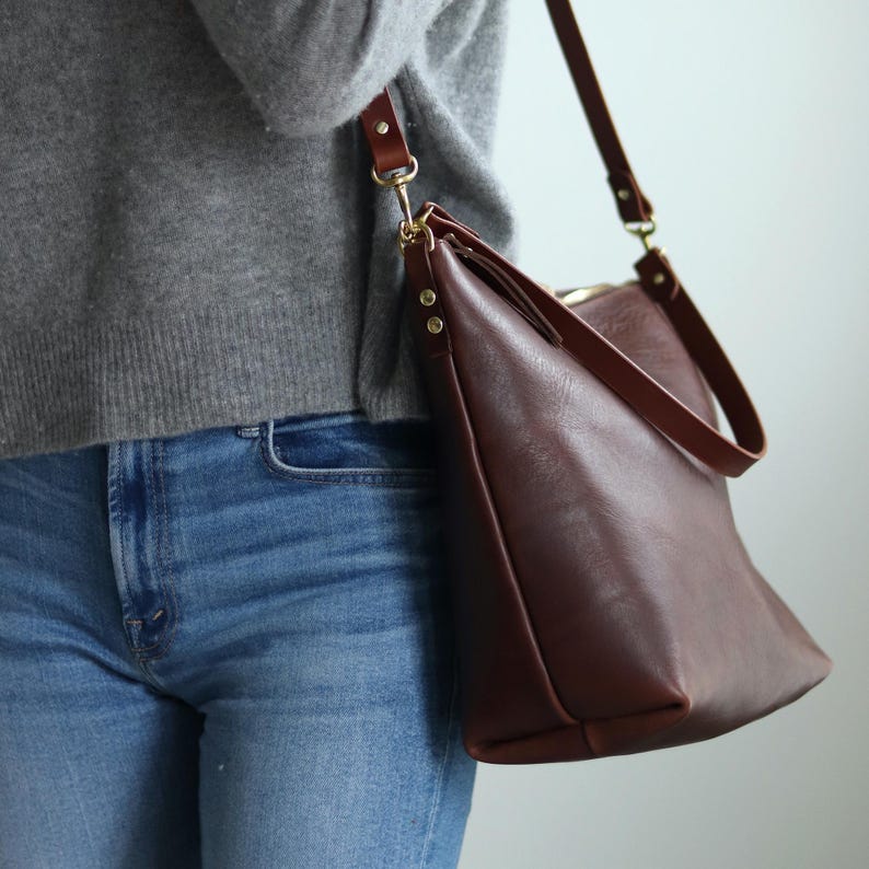Puede incluir: Un bolso de hombro de cuero marr&oacute;n con una correa larga. El bolso se lleva sobre el hombro de una persona que lleva un su&eacute;ter gris y jeans azules.