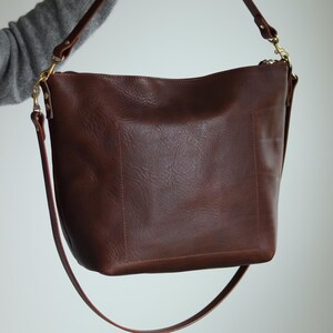 Puede incluir: Un bolso tote de cuero marr&oacute;n con una correa larga ajustable. El bolso tiene cierre de cremallera y un dise&ntilde;o sencillo.