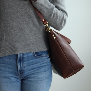 Puede incluir: Un bolso tote de cuero marr&oacute;n con una hebilla y una correa doradas. El bolso lo lleva una persona que lleva un su&eacute;ter gris y pantalones vaqueros azules.
