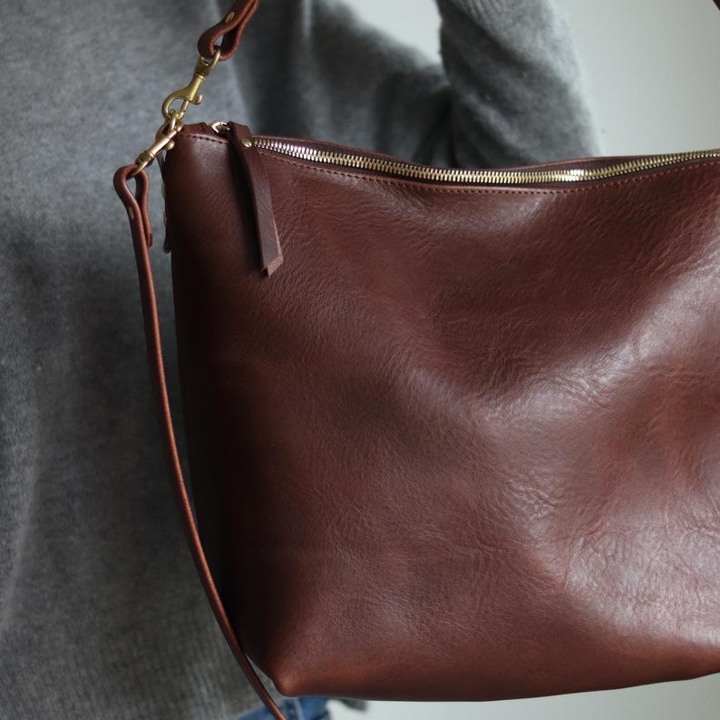 Puede incluir: Un bolso de hombro de cuero marr&oacute;n con cierre de cremallera y un tirador de cremallera dorado. El bolso tiene una correa larga que se puede ajustar para adaptarse a diferentes alturas.