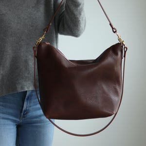 Puede incluir: Un bolso de hombro de cuero marr&oacute;n oscuro con una correa larga. El bolso tiene una cremallera y un estilo informal y holgado.