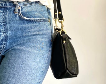 May include: A black leather crossbody bag with a gold buckle and strap. The bag is worn over the shoulder of a person wearing blue jeans.