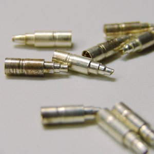 May include: A close-up of a group of silver-colored metal connectors. The connectors are small and cylindrical, with a slight taper at one end. They are arranged in a random pattern on a white surface.
