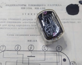 Może przedstawiać: Przezroczysta szklana rurka z różowym i srebrnym drutem w środku. Rurka jest oznaczona jako "ИН-12А" i stanowi część schematu obwodu z ponumerowanymi zaciskami. Schemat przedstawia punkty połączeń dla lampy wskaźnikowej z wyładowaniem jarzeniowym.