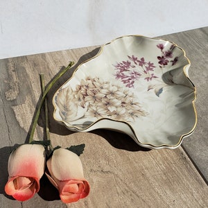 Może przedstawiać: Ozdobny ceramiczny talerz z falistym brzegiem i kwiatowym wzorem w odcieniach beżu, fioletu i złota. Dwie sztuczne róże z różowymi i czerwonymi płatkami obok talerza. Talerz ma złoty brzeg.