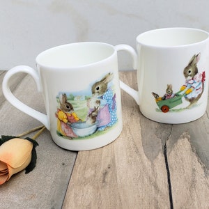 May include: Two white ceramic mugs with bunny illustrations. One mug features three bunnies washing clothes in a basin, while the other shows a bunny pulling a wagon with two other bunnies inside.