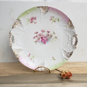 Może przedstawiać: Biała porcelanowa taca do serwowania z różowymi i zielonymi wzorami kwiatowymi i złotymi akcentami. Taca ma dwa uchwyty i falisty brzeg.