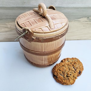 May include: A ceramic cookie jar shaped like a brown and tan barrel with a rope handle. The lid has a woven pattern. Two oatmeal cookies are in front of the jar.