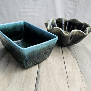 以下が含まれることがあります： 2つのセラミックプランター。1つは長方形で、ティール色で、葉の模様が施されています。もう1つは、濃い緑色の花型のボウルです。どちらも明るい木目調の表面に置かれています。