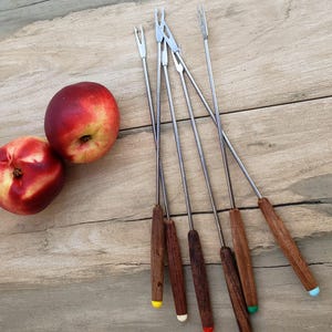 Peut inclure: Ensemble de fourchettes à fondue avec manches en bois et embouts colorés, à côté de deux nectarines mûres. Les fourchettes ont des dents en acier inoxydable et sont disposées sur une surface en bois.