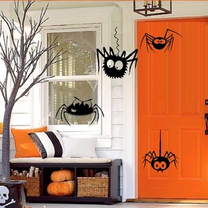 May include: A white door with a bright orange door, decorated with black spider cutouts. The door is surrounded by a white trim and a white wall. There is a window to the left of the door with a black spider cutout on the glass.
