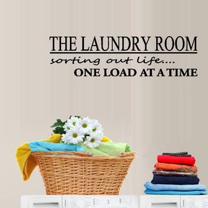 May include: A white laundry room wall decal with the text "The Laundry Room sorting out life... One Load At A Time". A wicker laundry basket with towels and flowers sits on top of a white washing machine.