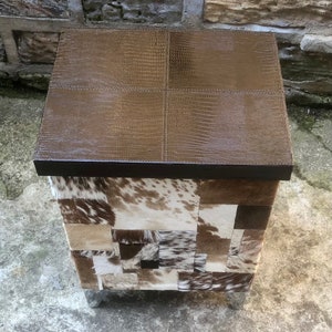 May include: A square ottoman with a brown leather top and a patchwork design of cowhide in shades of brown, white, and black. The ottoman has silver metal legs.
