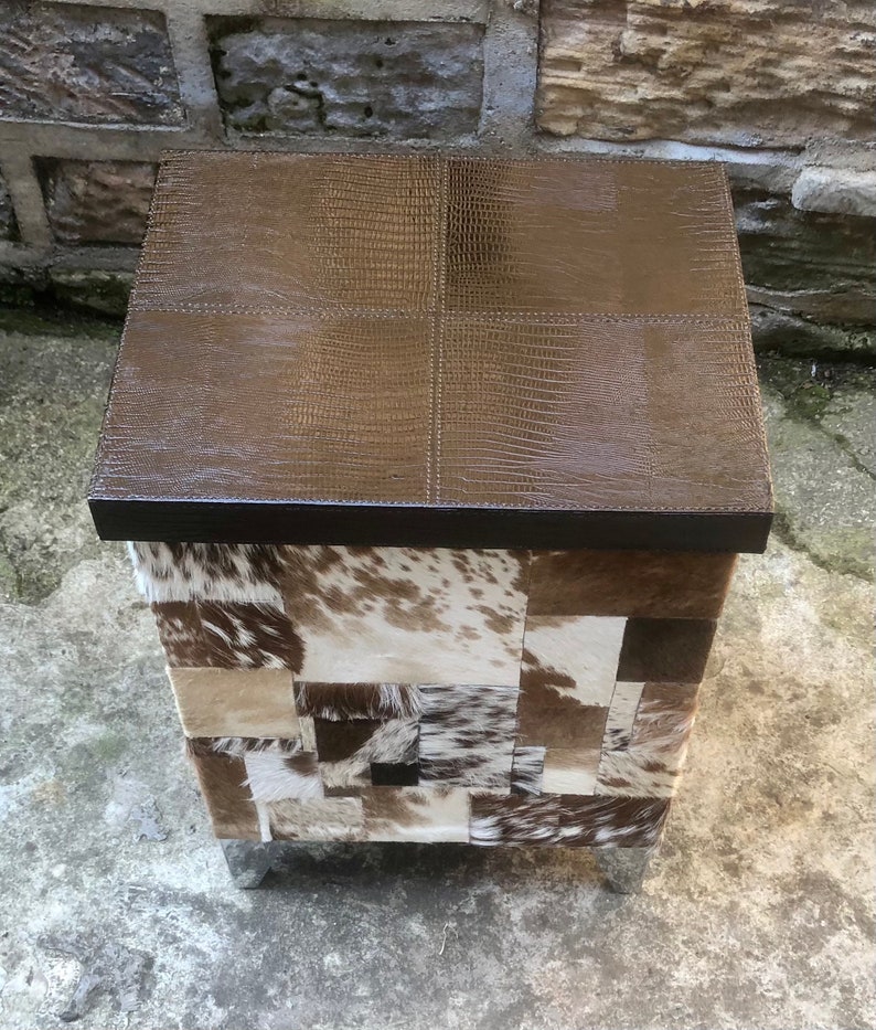 May include: A square ottoman with a brown leather top and a patchwork design of cowhide in shades of brown, white, and black. The ottoman has silver metal legs.