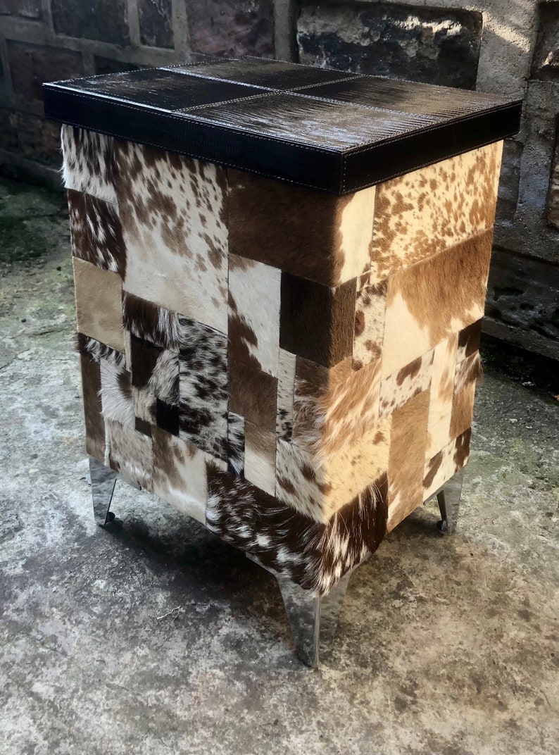 May include: A square storage ottoman with a patchwork design of brown, white and black cowhide. The ottoman has a black leather top and silver metal legs.