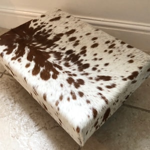 Puede incluir: Un reposapi&eacute;s rectangular cubierto de piel de vaca blanca y marr&oacute;n.