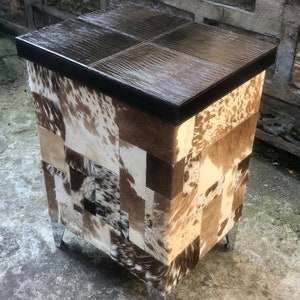 May include: A brown leather-topped storage ottoman with a patchwork design of cowhide in shades of brown, white, and black. The ottoman has four metal legs.