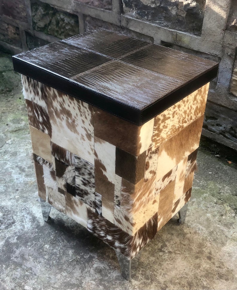 May include: A brown leather-topped storage ottoman with a patchwork design of cowhide in shades of brown, white, and black. The ottoman has four metal legs.