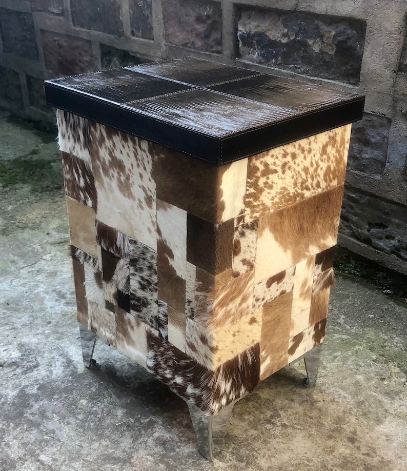 May include: A square, brown leather-topped ottoman with a patchwork design of cowhide in shades of brown, white, and tan. The ottoman has four silver metal legs.