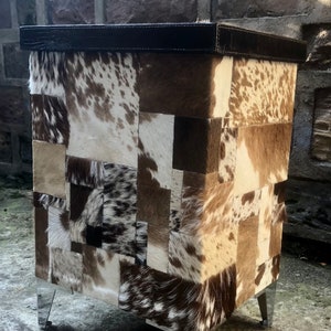 May include: A square ottoman with a patchwork design of brown, white and black cowhide. The ottoman has a dark brown leather top and silver metal legs.