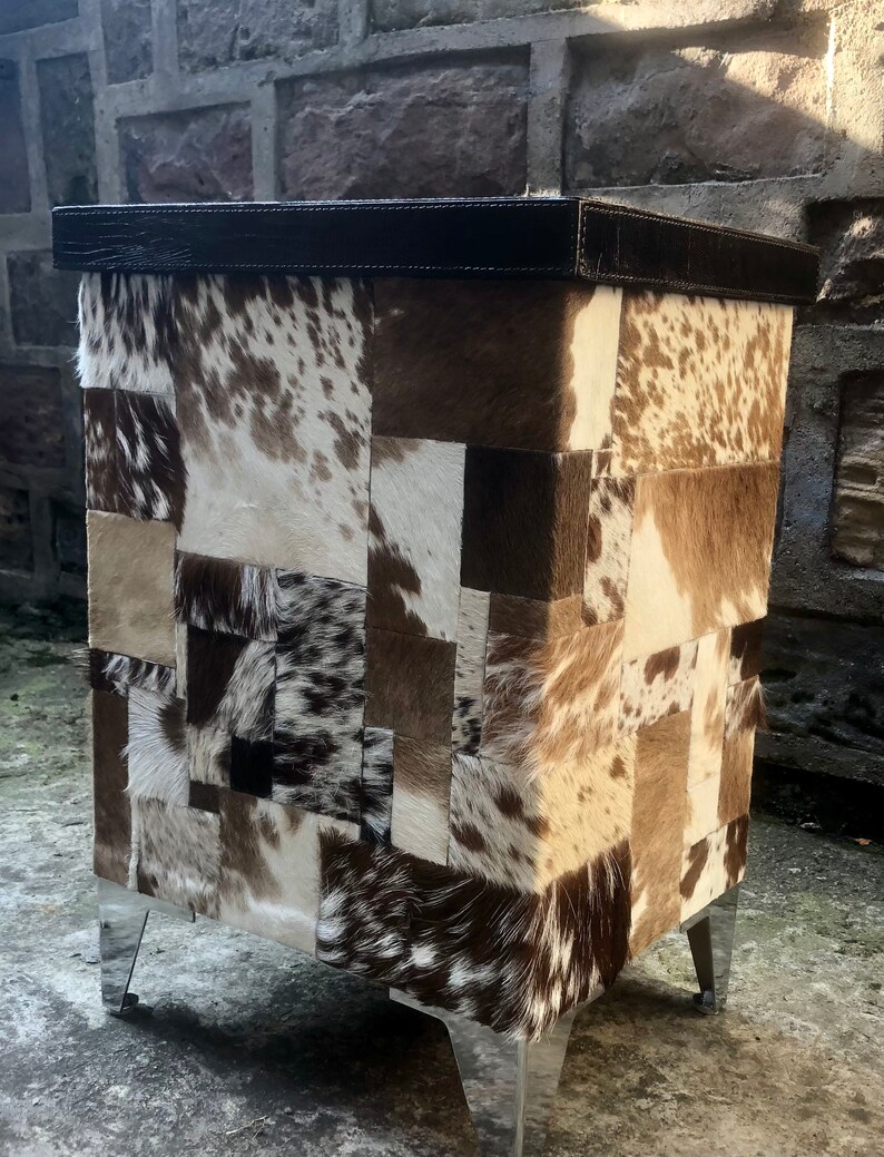 May include: A square ottoman with a patchwork design of brown, white and black cowhide. The ottoman has a dark brown leather top and silver metal legs.