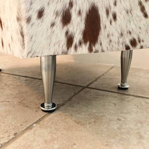 Puede incluir: Primer plano de un puf de piel de vaca blanca y marr&oacute;n con patas de metal plateado. El puf est&aacute; sobre un suelo de baldosas.