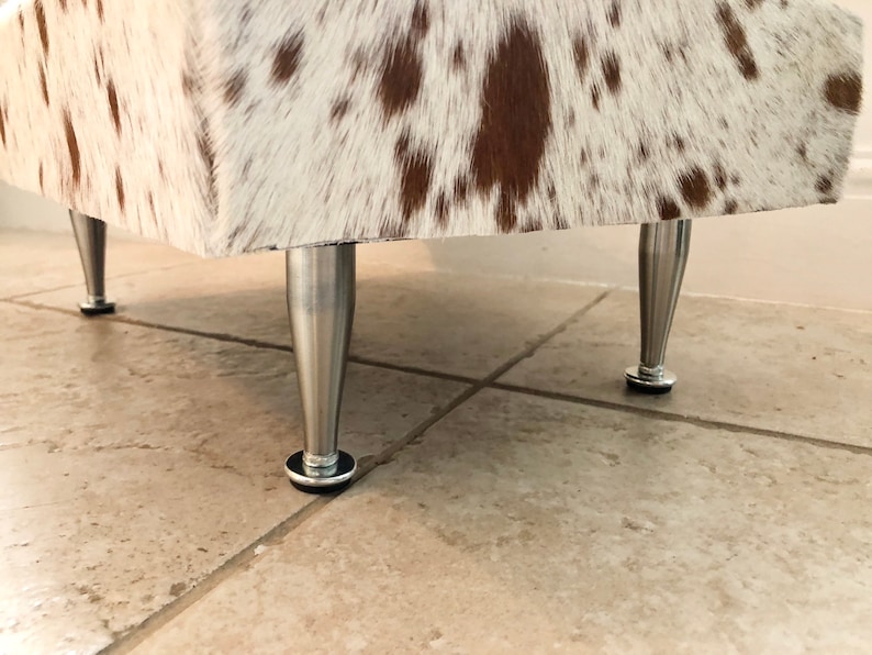 Puede incluir: Primer plano de un puf de piel de vaca blanca y marr&oacute;n con patas de metal plateado. El puf est&aacute; sobre un suelo de baldosas.