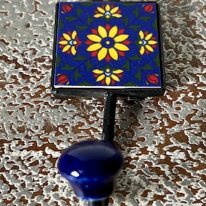Könnte beinhalten: Ein dekorativer Wandhaken mit quadratischer, blauer Fliesenoberseite mit einem Blumendesign in Gelb, Rot und Grün. Der Haken hat einen dunkelblauen, abgerundeten Knopf und einen schwarzen Metallschaft. Der Haken ist auf einer strukturierten, hellbraunen Oberfläche montiert.