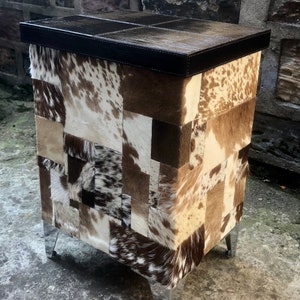 May include: A small, square, patchwork ottoman with a brown leather top and silver metal legs. The ottoman is covered in a variety of cowhide patches in shades of brown, white, and black.