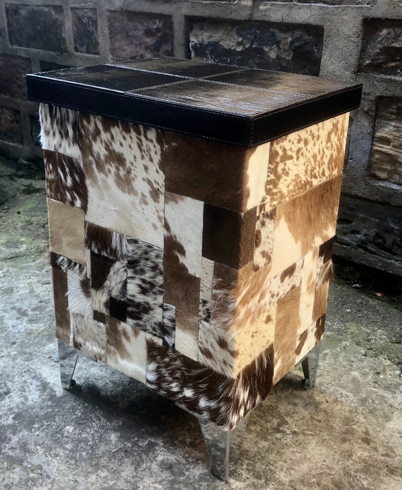 May include: A small, square, patchwork ottoman with a brown leather top and silver metal legs. The ottoman is covered in a variety of cowhide patches in shades of brown, white, and black.