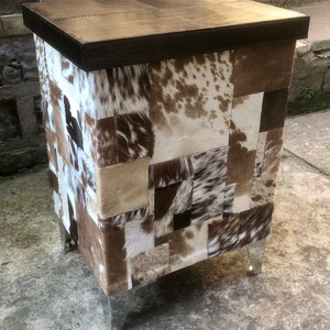 May include: A square-shaped, brown leather-topped stool with a patchwork design of brown, white, and black cowhide. The stool has four silver metal legs.