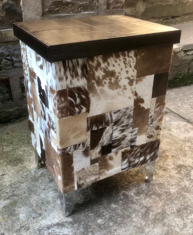 May include: A square-shaped, brown leather-topped stool with a patchwork design of brown, white, and black cowhide. The stool has four silver metal legs.